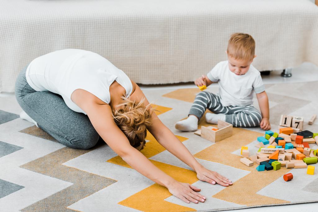 vrouw doet stretching oefening op tapijt en peuter jongen spelen met veelkleurige houten kubussen in woonkamer - Foto, afbeelding