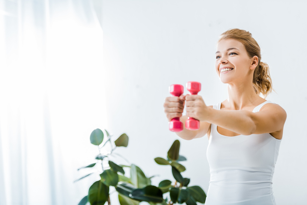 mulher atraente em sportswear sorrindo e fazendo exercício com halteres em casa
 - Foto, Imagem