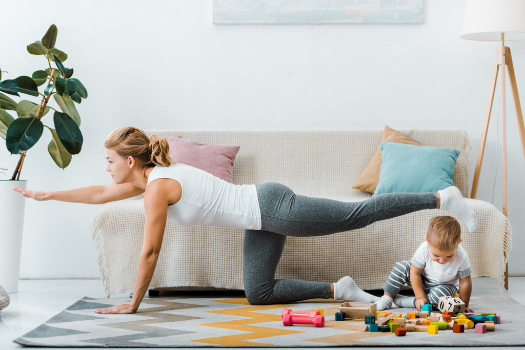 Kobieta, ćwiczenia w pobliżu kanapie i toddler chłopiec bawi się wielobarwny drewniane kostki na dywanie w salonie - Zdjęcie, obraz