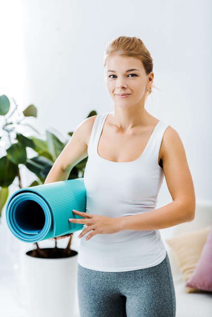 attraktive Frau in Sportbekleidung hält Fitnessmatte in der Hand und blickt in die Kamera im Wohnzimmer - Foto, Bild