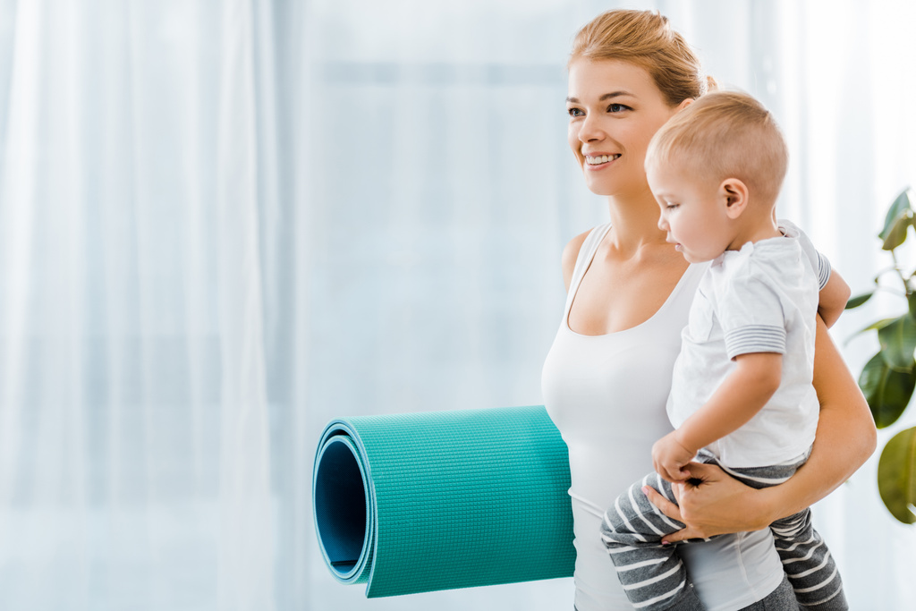 beautiful woman in sportswear holding blue fitness mat and smiling toddler boy - Photo, Image