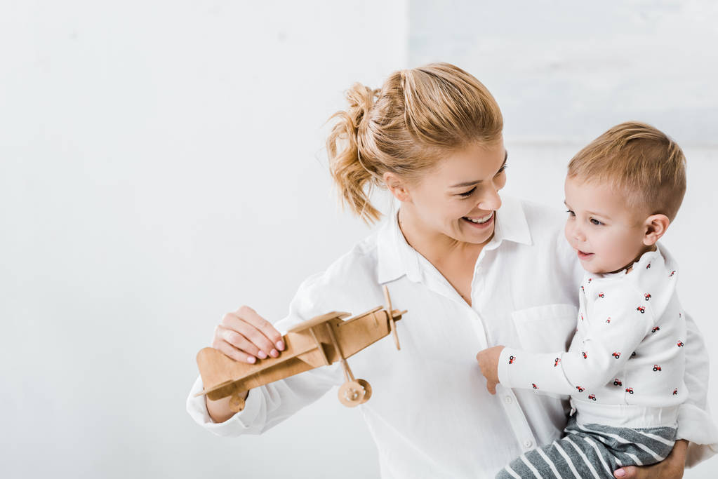 woman smiling, holding wooden plane model and playing with cute toddler boy at home - Photo, Image