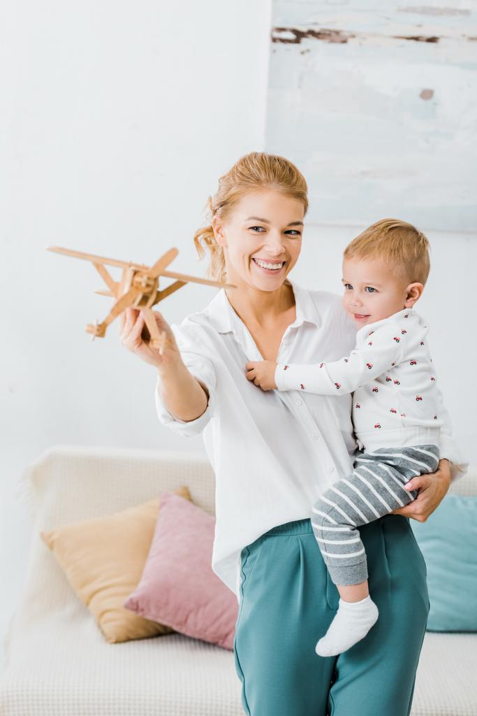 Lachende vrouw houden van houten vliegtuig model en spelen met schattige peuter jongen in woonkamer - Foto, afbeelding