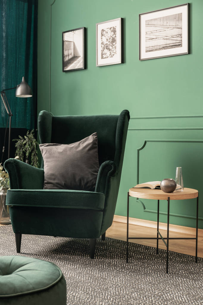 Серая подушка на изумрудно-зеленом кресле в зеленом интерьере гостиной с плакатами на стене и деревянным журнальным столиком
 - Фото, изображение