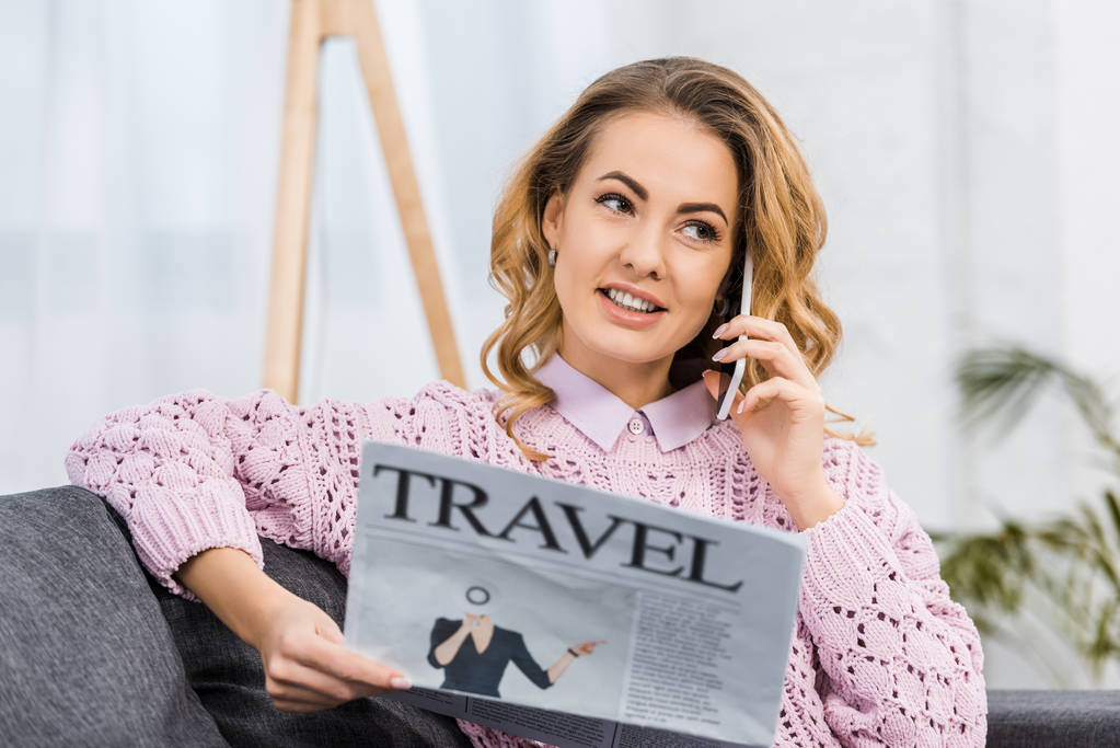 mooie vrouw zittend op de Bank, praten over smartphone en houden van reizen krant in woonkamer - Foto, afbeelding