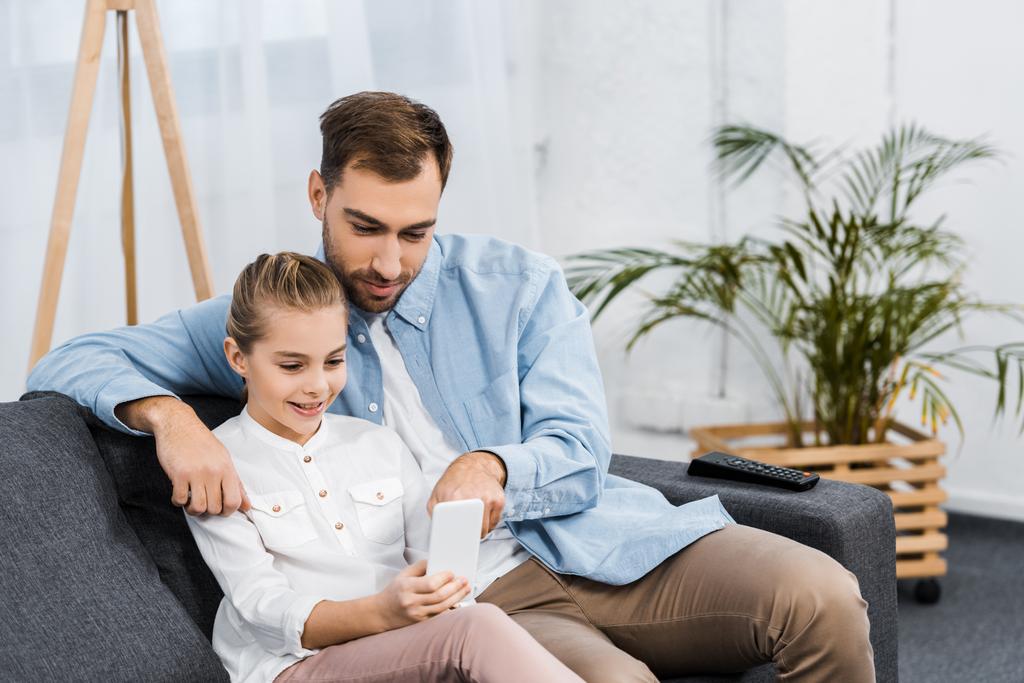 smiling daughter sitting on sofa and father pointing with finger at screen in apartment - Photo, Image