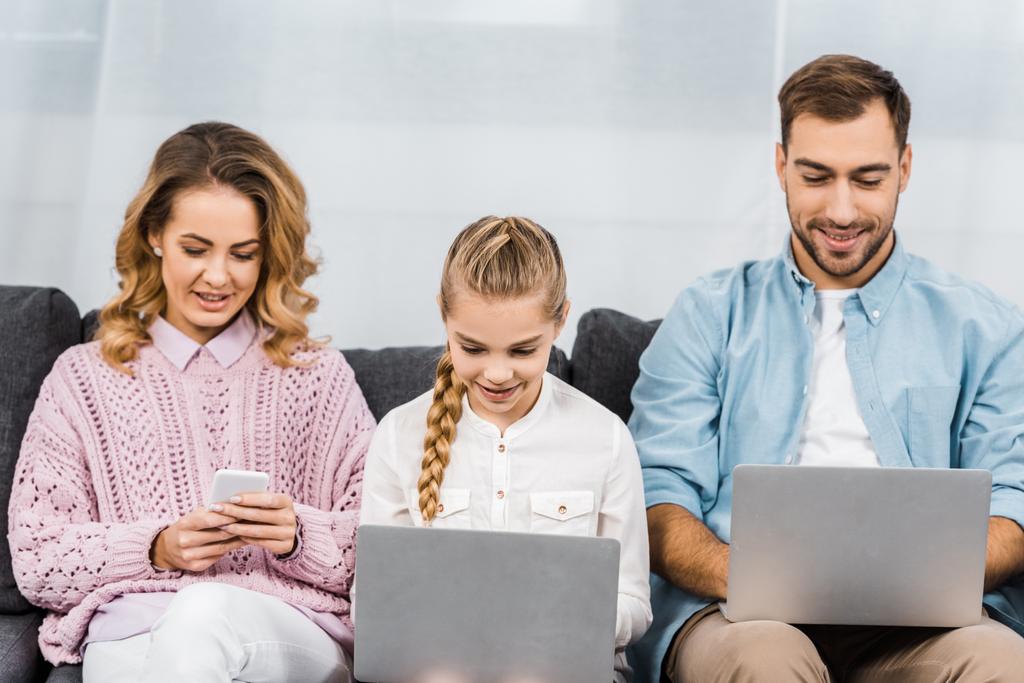 mooie vrouw zittend op de Bank en het gebruik van smartphone terwijl dochter en echtgenoot te typen op een toetsenbord van de laptop in de woonkamer - Foto, afbeelding