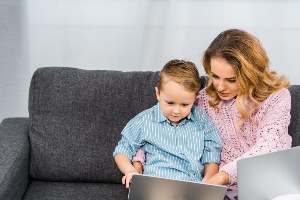 pretty woman with son sitting on sofa and pointing with finger on laptop screen in apartment - Photo, Image
