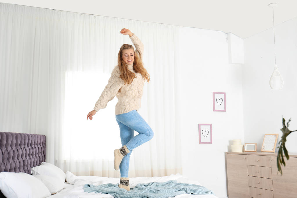 attraktive lächelnde junge Frau im kuschelig warmen Pullover, die zu Hause auf dem Bett springt - Foto, Bild