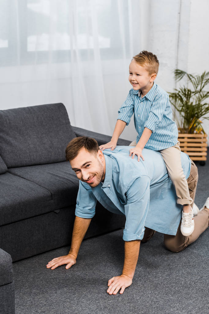 smiling boy sitting on back of father standing on all fours and looking at camera in living room - Photo, Image