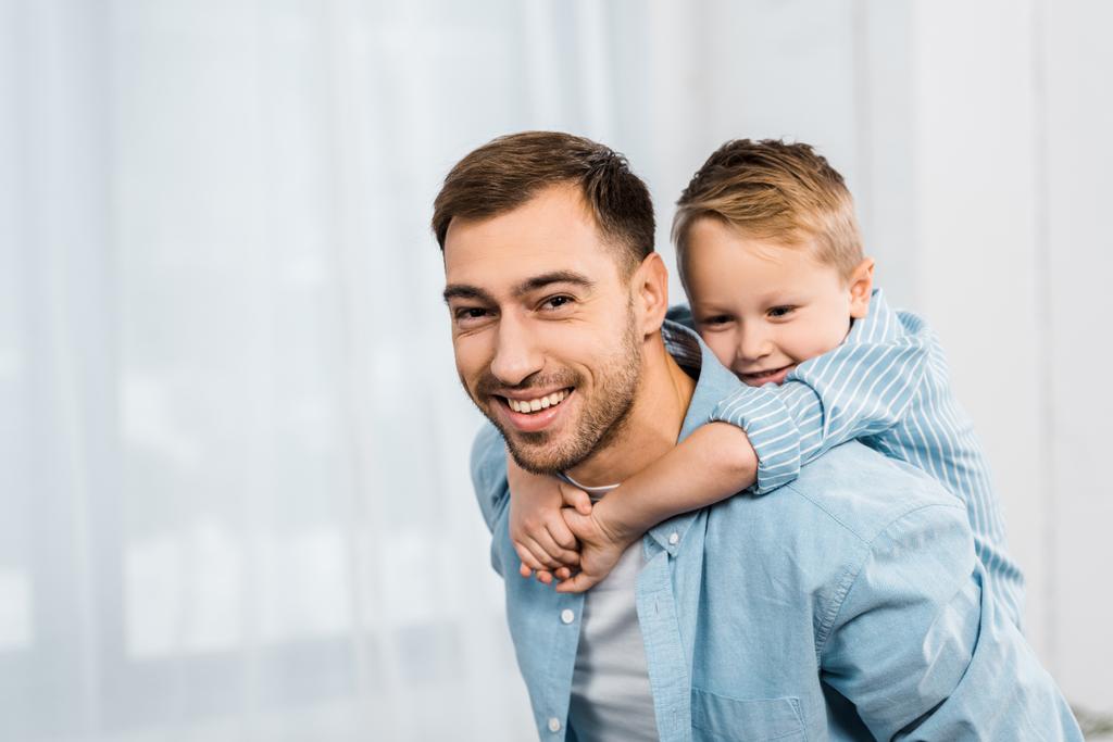 Улыбающийся отец держит милого сына на плечах и смотрит в камеру
 - Фото, изображение