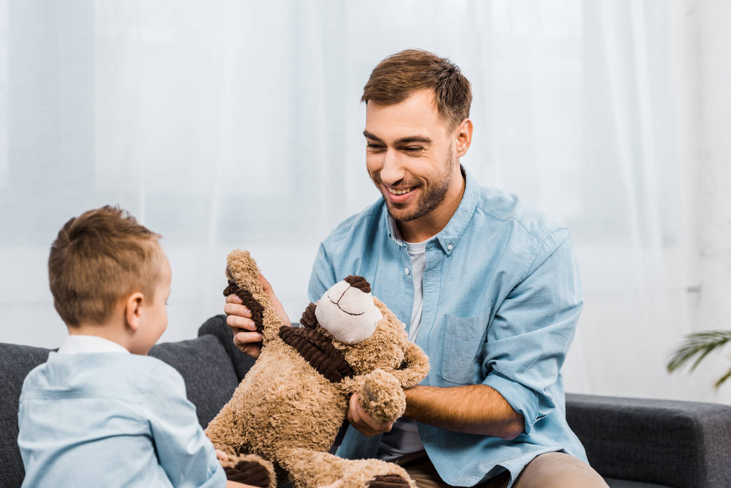 усміхнений батько і син сидять на дивані і тримають плюшевого ведмедя у вітальні
 - Фото, зображення