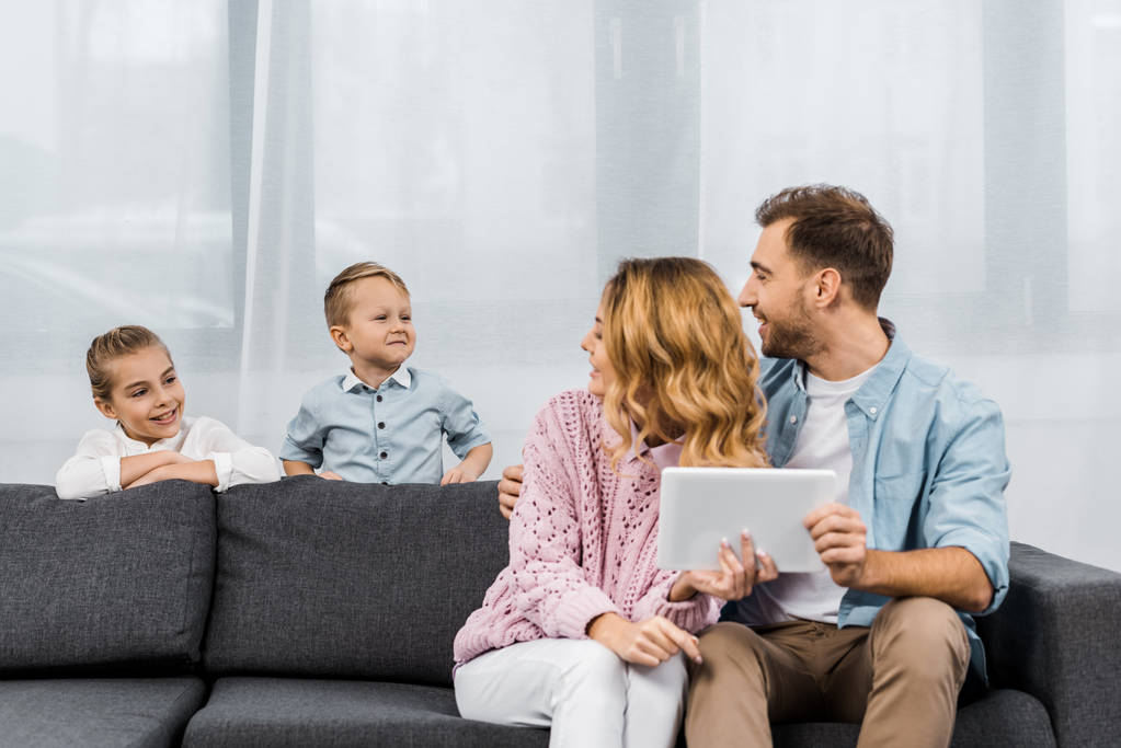 родители сидят на диване, держат цифровой планшет и смотрят на детей в гостиной
 - Фото, изображение