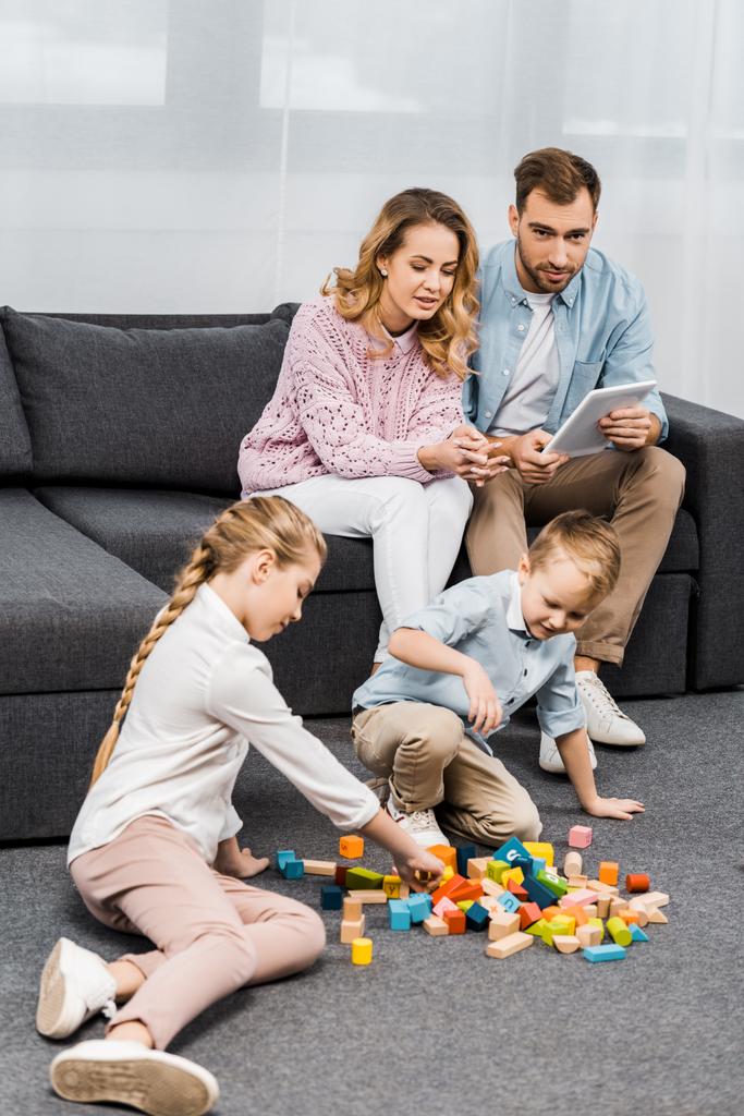 родители сидят на диване с цифровым планшетом и смотрят на детей, играющих с разноцветными деревянными блоками на полу в квартире
 - Фото, изображение