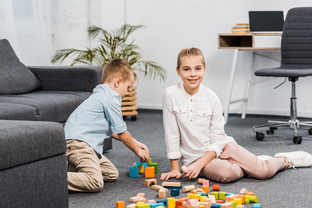 cute girl sitting on floor and looking at camera while boy  playing with multicolored wooden blocks in apartment - Photo, Image