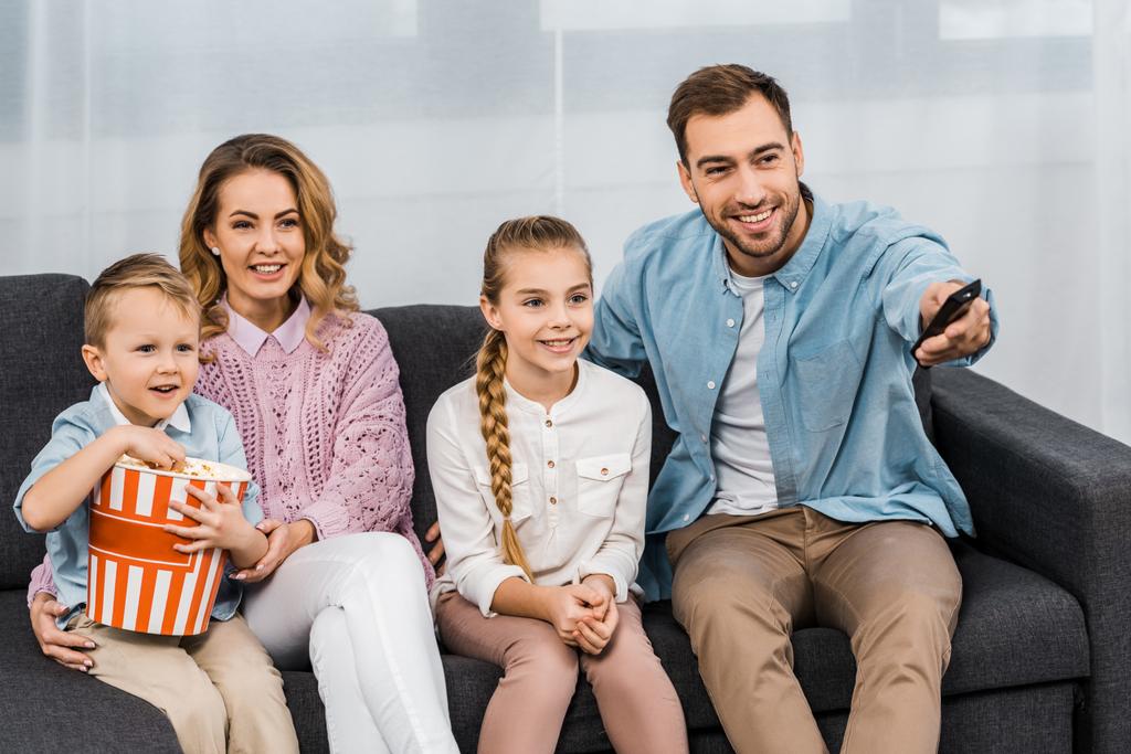 przystojny ojciec siedzi na kanapie i zmiana kanałów przez pilota z uśmiechający się żona i córka i syn jedzenie popcorn w mieszkaniu - Zdjęcie, obraz