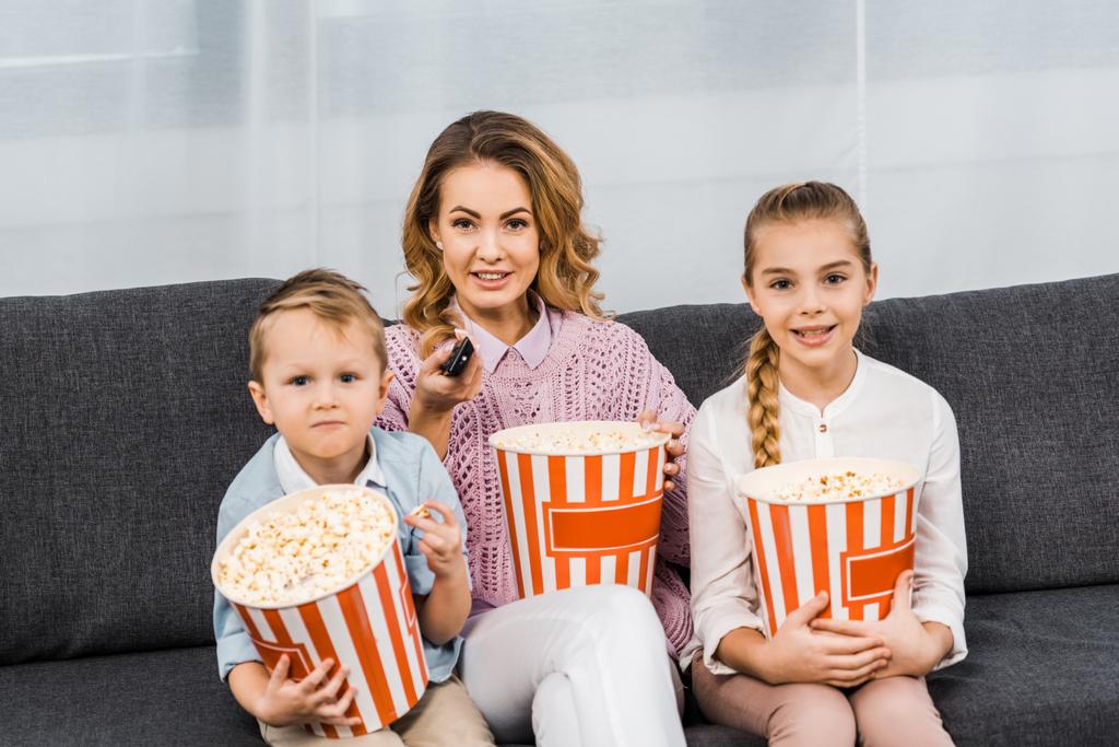 улыбающаяся мать с детьми, сидящими на диване, держащими полосатые ведра попкорна и переключающими каналы пульта дистанционного управления в гостиной
 - Фото, изображение