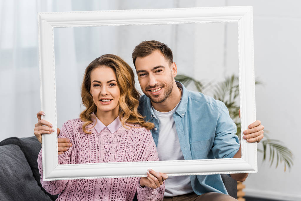 sonriente hombre y mujer sosteniendo marco blanco y mirando a la cámara en la sala de estar
 - Foto, Imagen