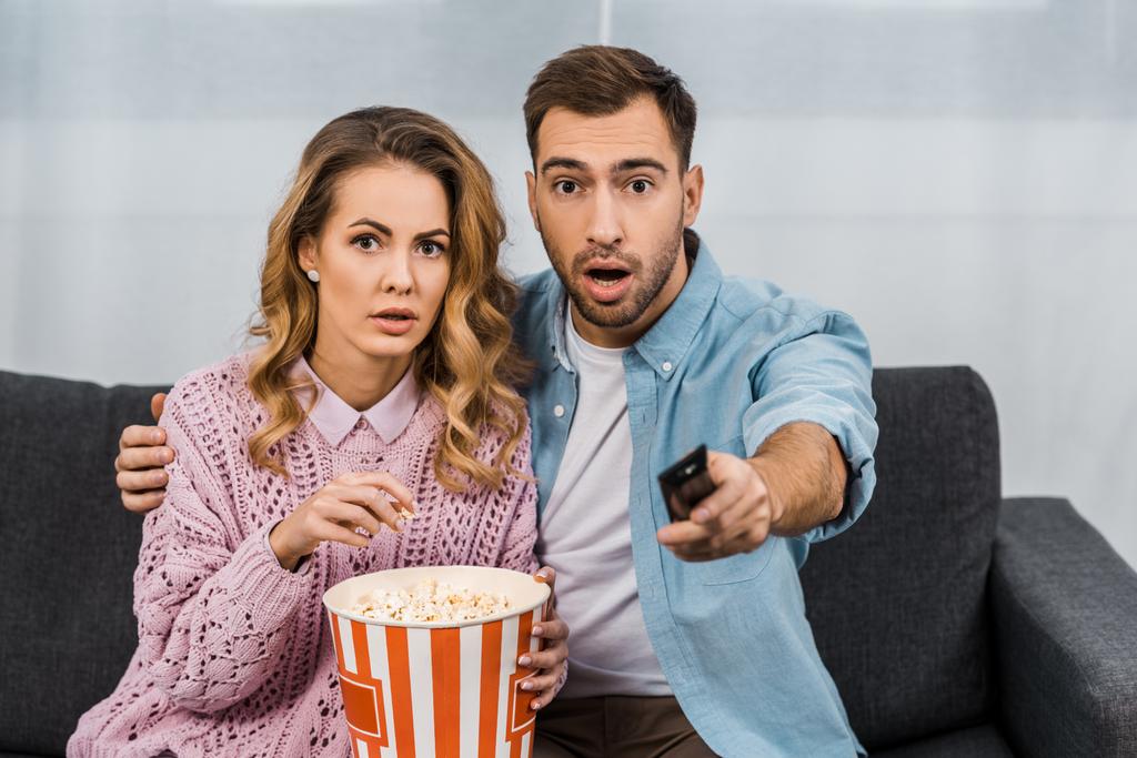 open-eyed man sitting on sofa, holding remote controller and embracing surprised wife with striped popcorn bucket - Photo, Image