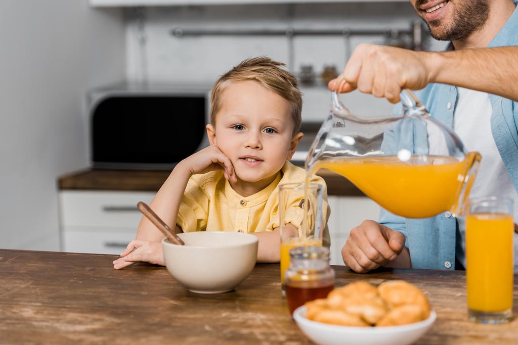 обрізаний вид усміхненого батька, що лив апельсиновий сік у склянку і милий хлопчик дивиться на камеру
 - Фото, зображення