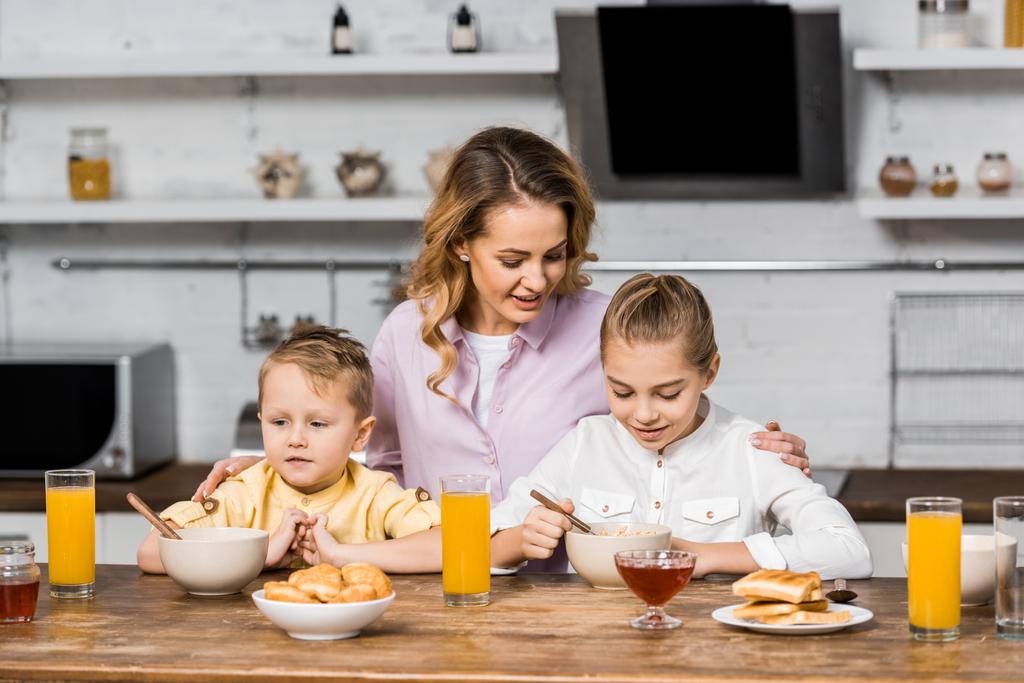 mulher bonita abraçando crianças bonitos comer aveia na mesa da cozinha
 - Foto, Imagem