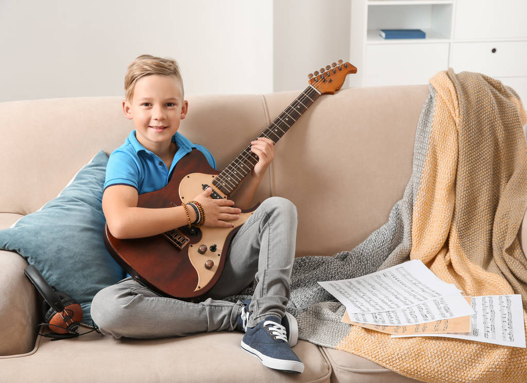 Милый мальчик играет на гитаре на диване в комнате. Пространство для текста
 - Фото, изображение