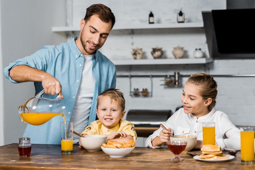 sonriente padre vertiendo jugo de naranja en un vaso mientras los niños comen gachas en la mesa
 - Foto, Imagen