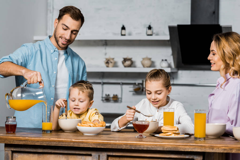 όμορφος ο πατέρας ρίχνει χυμός πορτοκαλιού στο ποτήρι, ενώ τα παιδιά που τρώνε πλιγούρι βρώμης στο τραπέζι - Φωτογραφία, εικόνα