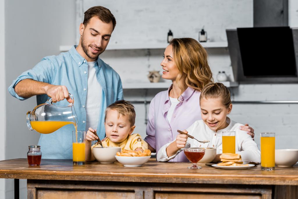 Lächelnde Frau schaut Ehemann an, der Orangensaft ins Glas gießt, während Kinder Haferflocken essen - Foto, Bild