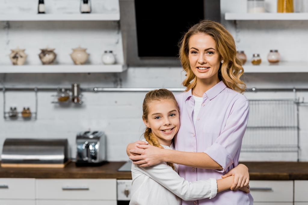 aantrekkelijke vrouw en schattige dochter knuffelen en camera in de keuken kijken - Foto, afbeelding