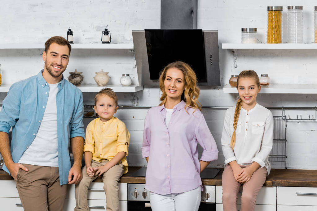 улыбающиеся родители с дочерью и сыном смотрят в камеру на кухне
 - Фото, изображение