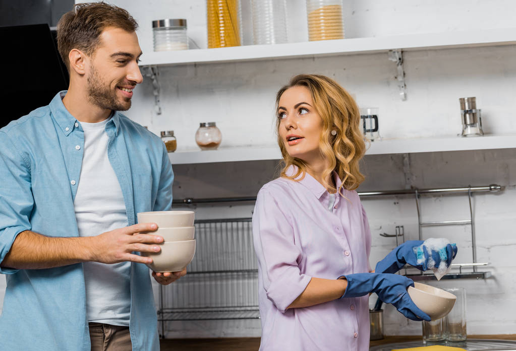 привлекательная женщина в резиновых перчатках моет посуду и смотрит на улыбающегося мужа, держащего миски на кухне
 - Фото, изображение
