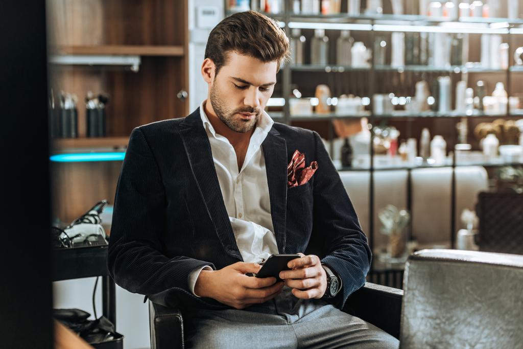 élégant beau jeune homme en utilisant smartphone tout en étant assis dans un salon de beauté
 - Photo, image