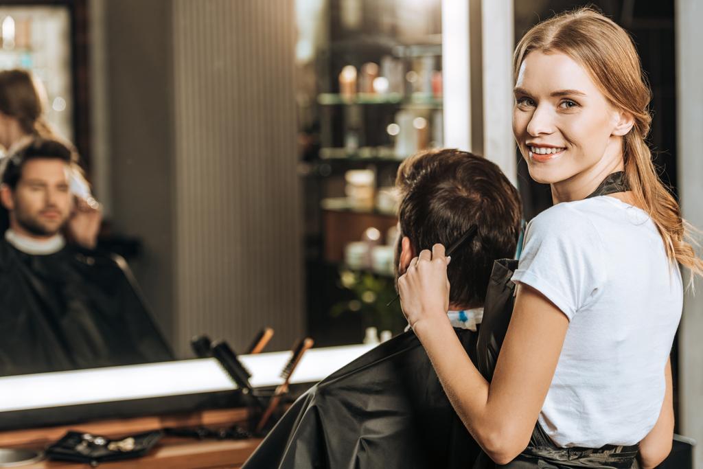 beautiful young hairstylist smiling at camera while combing hair to male client in beauty salon  - Photo, Image