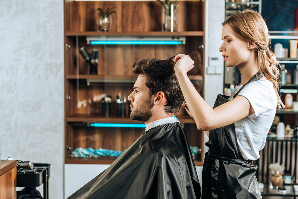 Seitenansicht des Friseurs, der einem hübschen jungen Mann im Schönheitssalon die Haare schneidet   - Foto, Bild