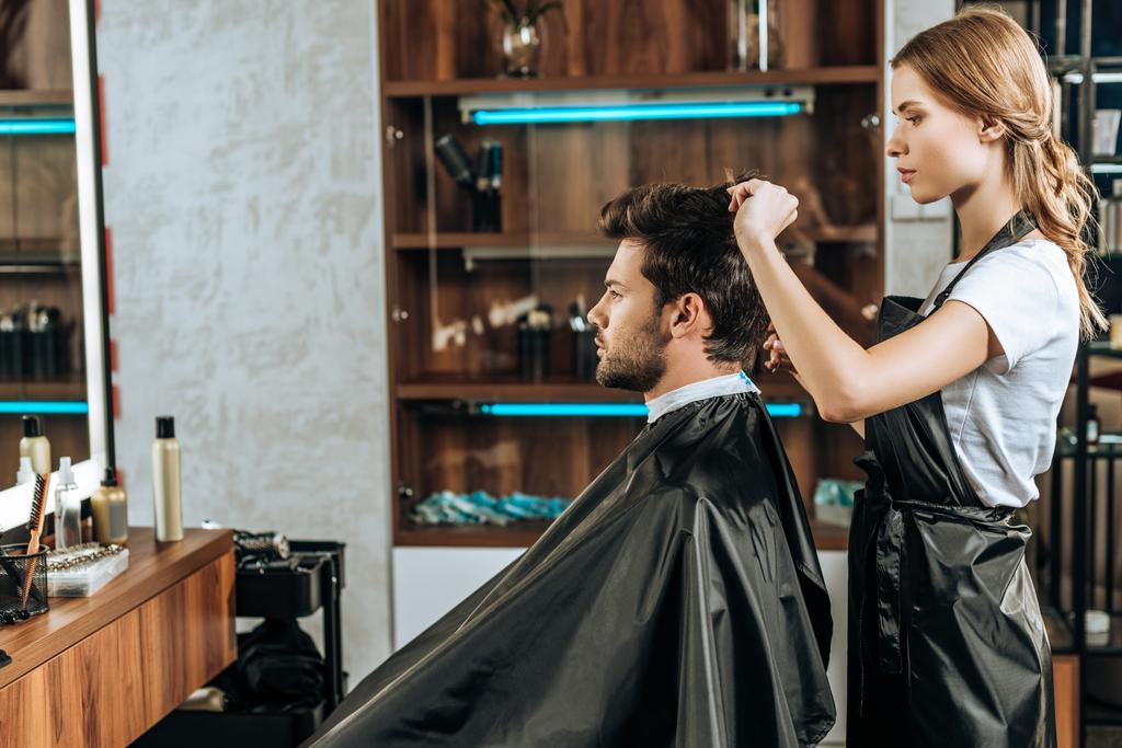 side view of hairstylist cutting hair to handsome young man in beauty salon   - Photo, Image