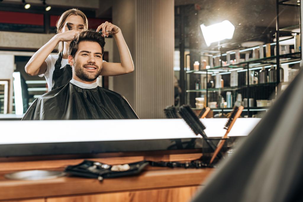 reflection in mirror of young hairdresser cutting hair to handsome smiling man in beauty salon - Photo, Image