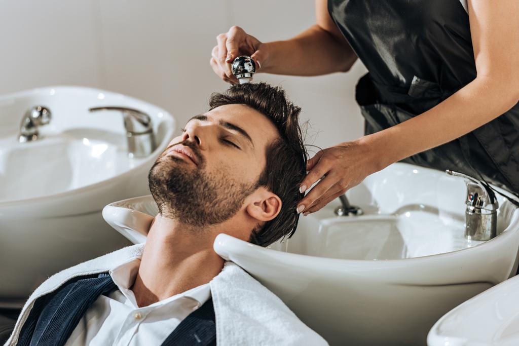 abgeschnittene Aufnahme eines jungen Friseurs, der einem hübschen Kunden im Schönheitssalon die Haare wäscht - Foto, Bild