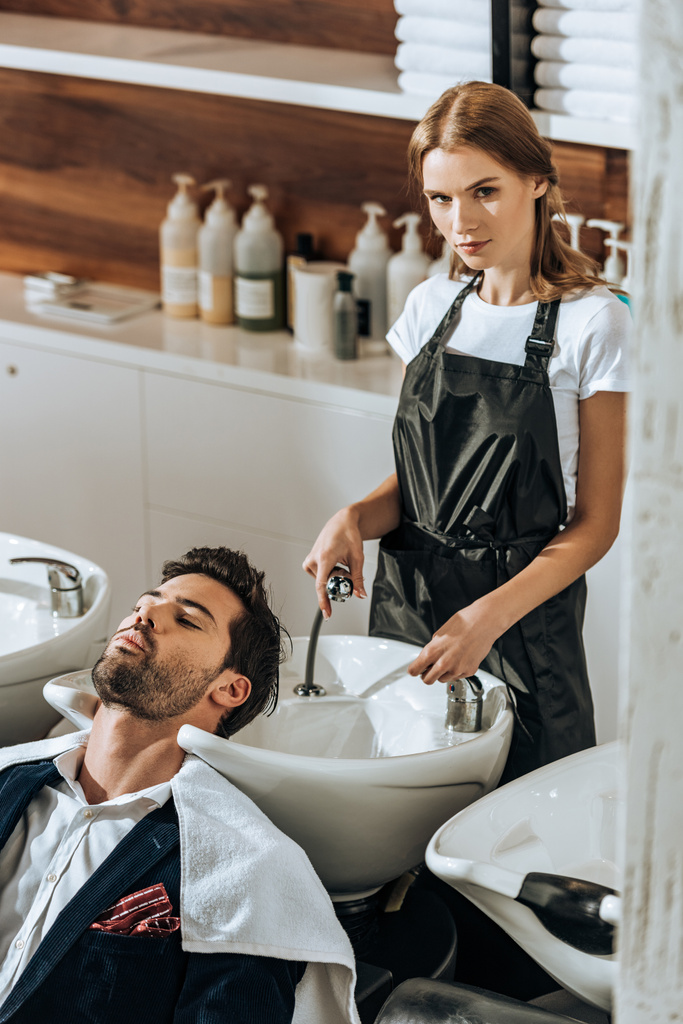 vista de ángulo alto de peluquero mirando a la cámara mientras lava el cabello a un joven guapo en el salón de belleza
  - Foto, imagen