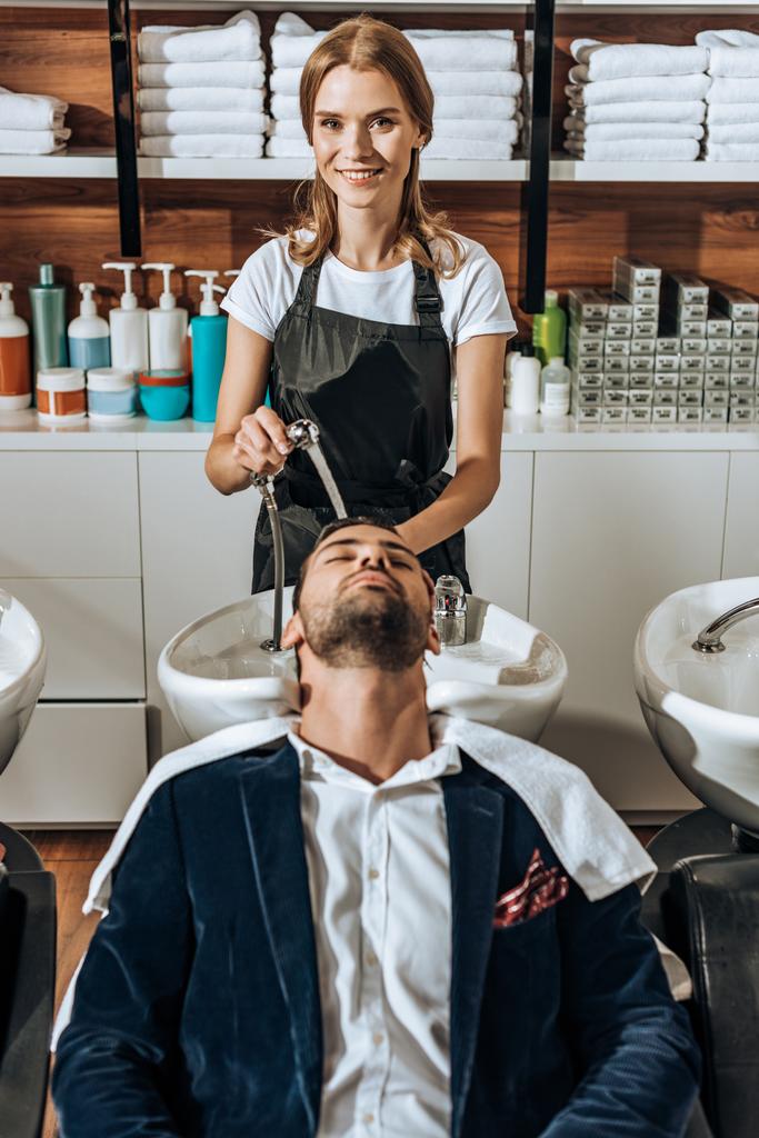 schöne junge Friseurin lächelt in die Kamera, während sie einem hübschen Mann im Schönheitssalon die Haare wäscht - Foto, Bild