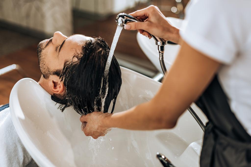 ビューティー サロンでハンサムな若い男に髪を洗うのヘアスタイリストの部分的なビュー   - 写真・画像