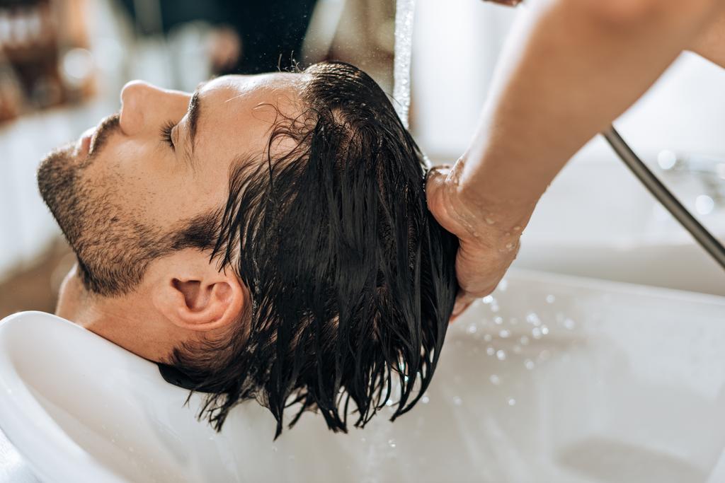 abgeschnittene Aufnahme eines Friseurs, der einem hübschen jungen Mann im Schönheitssalon die Haare wäscht   - Foto, Bild