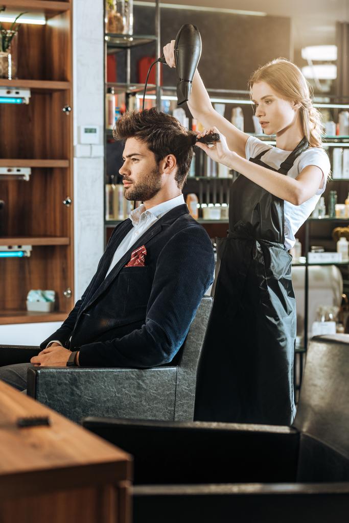 vista lateral del hermoso peinado joven peinando y secando el cabello al cliente guapo en el salón de belleza
 - Foto, Imagen