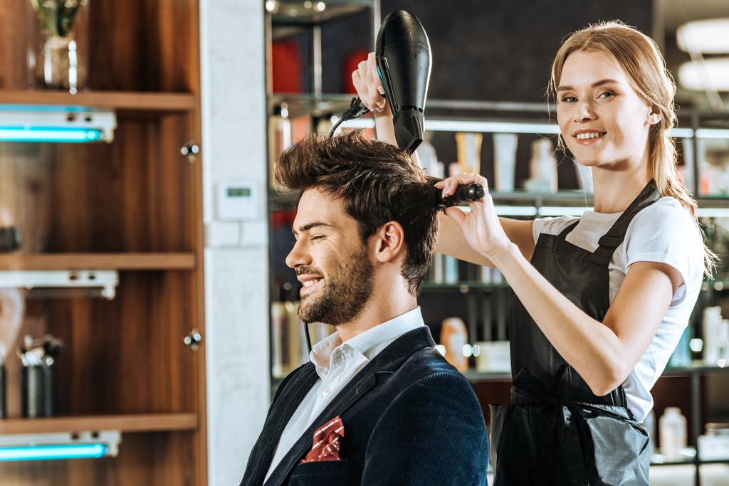 beautiful young hairstylist smiling at camera while drying and combing hair to handsome client in beauty salon - Photo, Image