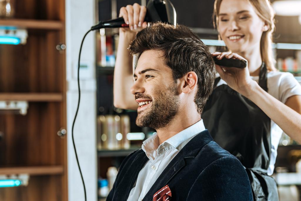 smiling young hairstylist drying hair to handsome happy man in beauty salon - Photo, Image