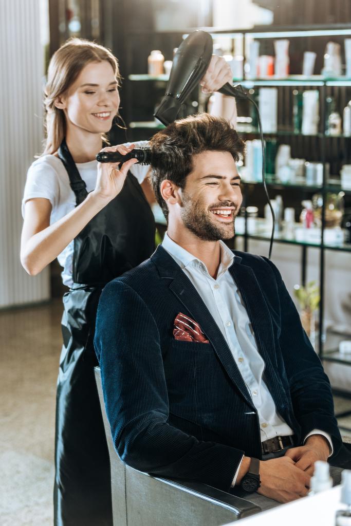 young hairstylist combing and drying hair to smiling handsome client in beauty salon  - Photo, Image