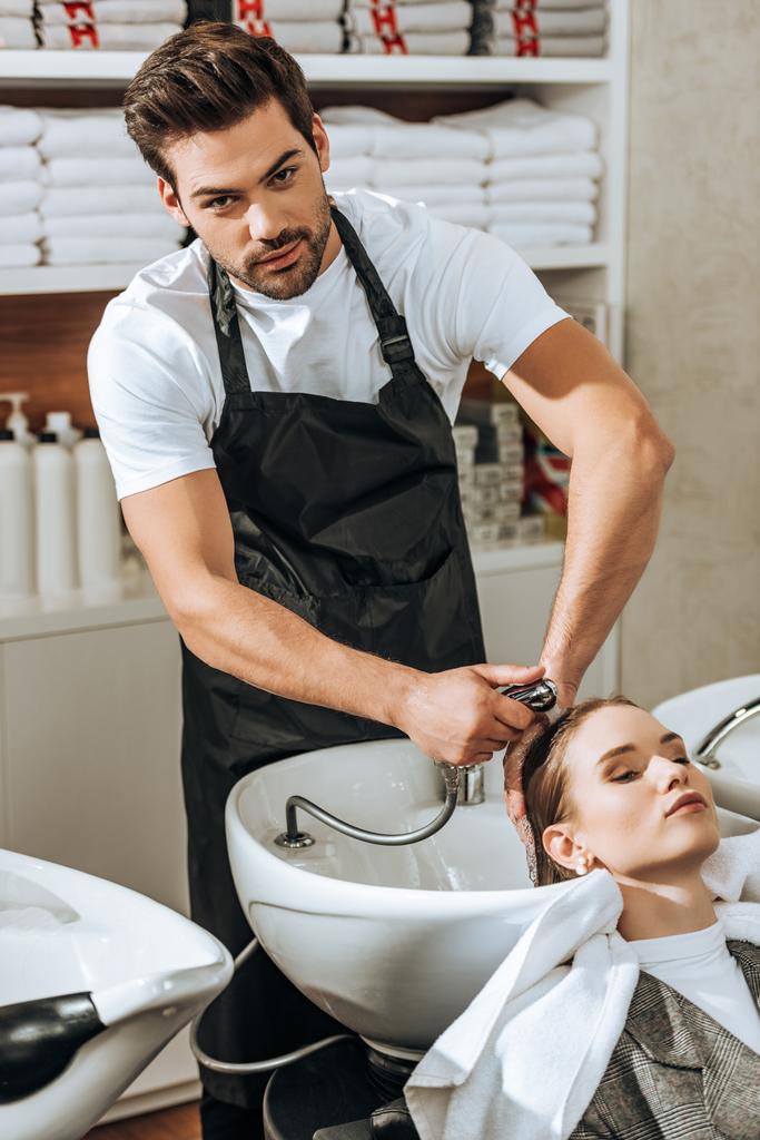 pohledný mladý kadeřník při mytí vlasy s ženou v salonu krásy při pohledu na fotoaparát  - Fotografie, Obrázek