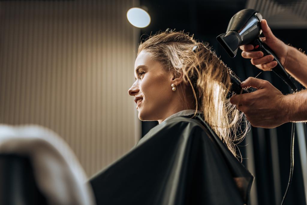 Oříznout záběr kadeřník sušení vlasů na usmívající se dívka v salonu krásy  - Fotografie, Obrázek