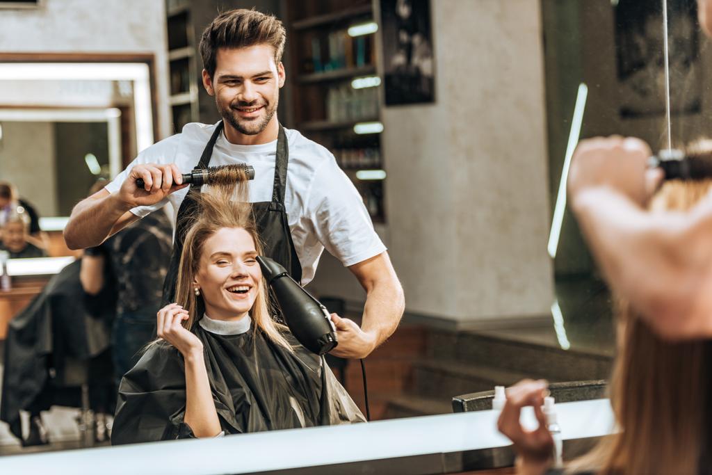 улыбающийся молодой парикмахер расчесывает и сушит волосы счастливой молодой женщине в салоне красоты
 - Фото, изображение