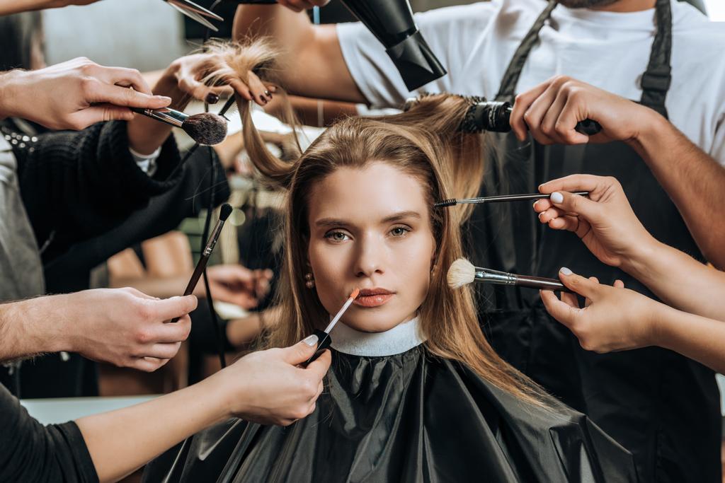 menina bonita olhando para a câmera enquanto estilistas aplicando maquiagem e fazendo penteado no salão de beleza
   - Foto, Imagem